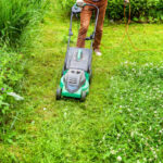 Man cutting green grass with lawn mower in backyard. Gardening country lifestyle background. Beautiful view on fresh green grass lawn in sunlight, garden landscape in spring or summer season