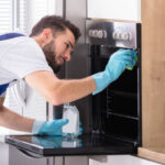 Happy Male Janitor Cleaning Oven With Sponge In Kitchen